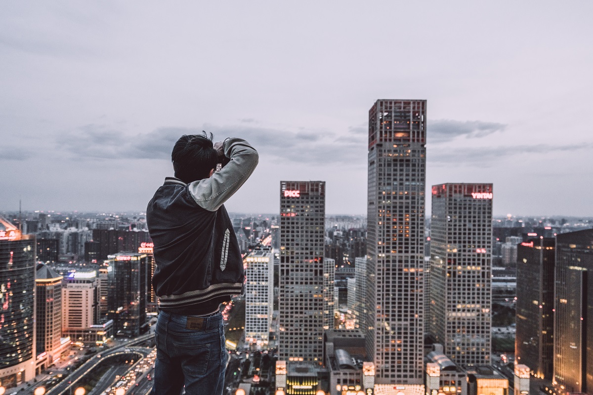 Hombre parado en una colina, sacando una foto a una ciudad muy moderna de China. Se ven edificios altos y está anocheciendo. El hombre está de espaldas.
