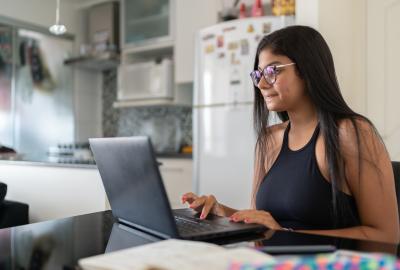 La imagen muestra a una joven mujer de cabello largo y lentes, frente a una computadora, estudiando en  la cocina de su casa