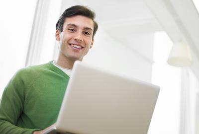 La imagen muestra a un joven sonriente mirando a cámara y sosteniendo una computadora abierta sobre sus piernas