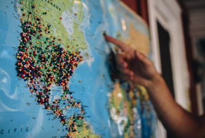 En la imagen se ve un mapa planisferio colgado en la pared, una mano que señana con el dedo y el mapa está marcado con muchas alfileres de colores