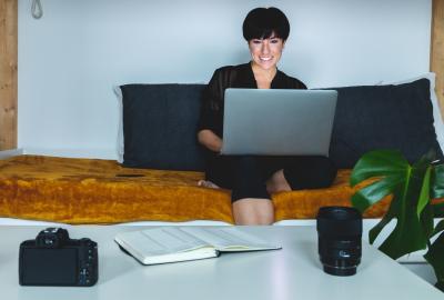 En la imagen se muestra a una mujer de pelo corto sentada en un sofá con la notebook, está sonriendo