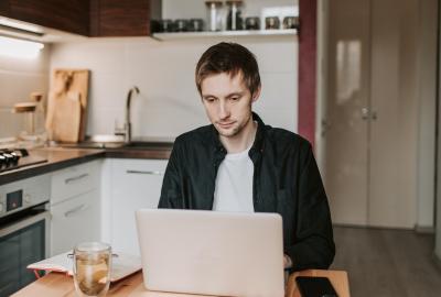 En la imagen se ve a un muchacho sentado a la mesa de la cocina, mirando una computadora.