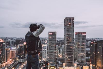 Hombre parado en una colina, sacando una foto a una ciudad muy moderna de China. Se ven edificios altos y está anocheciendo. El hombre está de espaldas.