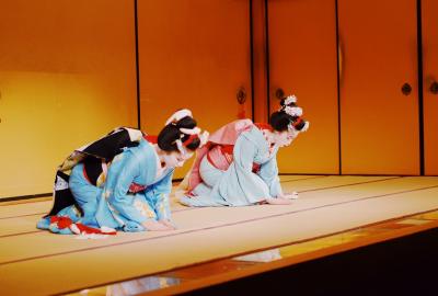 Dos geishas sobre un escenario, están agachadas de rodillas en señal de rezo