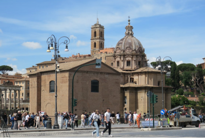La imagen muestra una foto de Roma donde se ve una iglesia y gente caminando por la calle