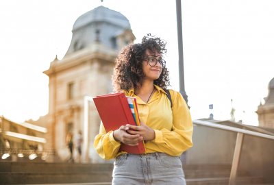 La imagen muestra a una estudiante sosteniendo un cuaderno y sonriendo, en la calle