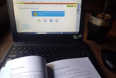 La imagen muestra un esciritorio con una computadora encendida, un libro con el texto de la fábula y un mate