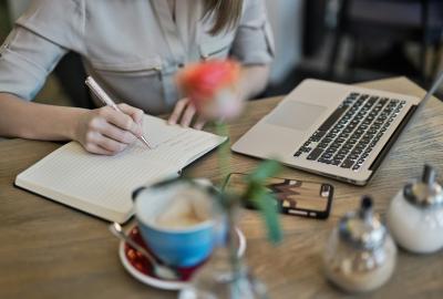 La imagen muestra un escritorio con una computadora, una taza y una persona escribiendo sobre un cuaderno